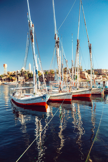 View of traditional feluccas docked on the Nile River with people on the tall masts and reflections in the calm water under a clear blue sky in Aswan Aswan Governorate Egypt. View of traditional feluccas docked on the Nile River with people on the tall masts and reflections in the calm water under a clear blue sky in Aswan Aswan Governorate Egypt.