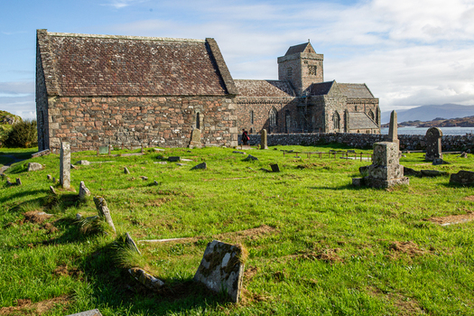 View of Iona Abbey and Nunnery with ancient stone headstones in a grassy graveyard under a bright sky Isle of Iona Scotland United Kingdom. View of Iona Abbey and Nunnery with ancient stone headstones in a grassy graveyard under a bright sky Isle of Iona Scotland United Kingdom.