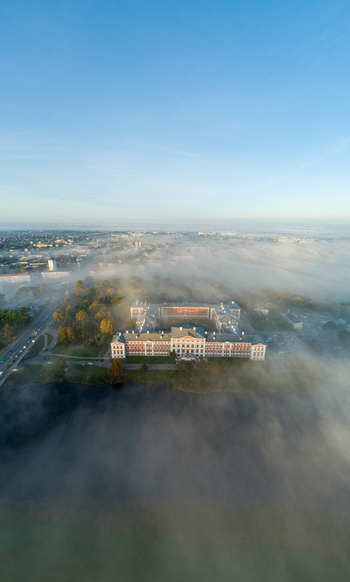 Aerial view of Jelgava Palace surrounded by dense morning fog with the Lielupe River in the foreground and clear blue sky above in Jelgava Latvia. Aerial view of Jelgava Palace surrounded by dense morning fog with the Lielupe River in the foreground and clear blue sky above in Jelgava Latvia.