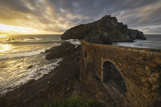 View of the historic stone bridge leading to San Juan de Gaztelugatxe hermitage under a dramatic sunset sky with crashing waves Gaztelugatxeko Doniene Euskadi Spain. View of the historic stone bridge leading to San Juan de Gaztelugatxe hermitage under a dramatic sunset sky with crashing waves Gaztelugatxeko Doniene Euskadi Spain.