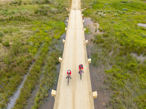 Aerial view of two cyclists riding across a concrete bridge surrounded by lush green marshland in Greyton Western Cape South Africa.