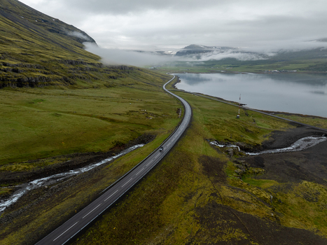 Aerial view of a winding road traversing the green landscape of the Eastern Region in Iceland with misty mountains a calm fjord and dramatic clouds Iceland. Aerial view of a winding road traversing the green landscape of the Eastern Region in Iceland with misty mountains a calm fjord and dramatic clouds Iceland.