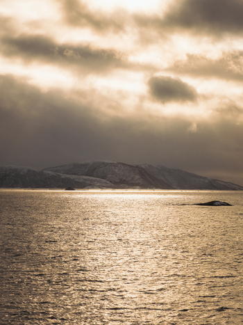 View of golden sunlight reflecting across the serene water towards the distant snowy mountains under a cloudy sky Brensholmen Troms Norway. View of golden sunlight reflecting across the serene water towards the distant snowy mountains under a cloudy sky Brensholmen Troms Norway.