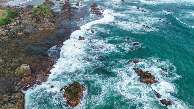 Aerial view of the rocky coastline with turquoise waves crashing against jagged rocks and dark volcanic formations in Indonesia. Aerial view of the rocky coastline with turquoise waves crashing against jagged rocks and dark volcanic formations in Indonesia.