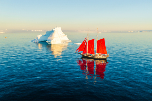 Aerial view of a sailboat with vibrant red sails gliding through calm blue waters past a massive iceberg in Greenland. Aerial view of a sailboat with vibrant red sails gliding through calm blue waters past a massive iceberg in Greenland.