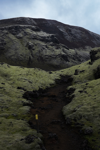 View of a dark path winding through mossy green hills under a cloudy sky leading towards a rugged mountain in Skaftárhreppur Iceland. View of a dark path winding through mossy green hills under a cloudy sky leading towards a rugged mountain in Skaftárhreppur Iceland.