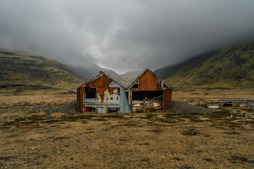 View of the old rusty corrugated metal huts at Stokksnes set against a backdrop of moss covered mountains under a heavy overcast sky in Iceland.