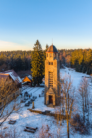 Aerial view of Bramberk lookout tower surrounded by snow covered forest and wooden buildings during golden hour in Lucany nad Nisou Liberec Region Czechia. Aerial view of Bramberk lookout tower surrounded by snow covered forest and wooden buildings during golden hour in Lucany nad Nisou Liberec Region Czechia.