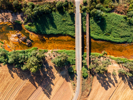 Aerial view of a bridge crossing a river with orange tinted water surrounded by lush green vegetation and golden agricultural fields in Andalusia Spain. Aerial view of a bridge crossing a river with orange tinted water surrounded by lush green vegetation and golden agricultural fields in Andalusia Spain.