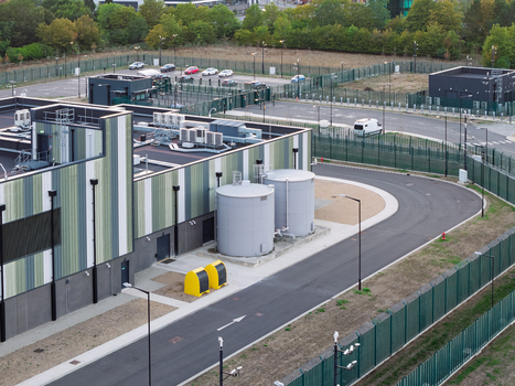 Aerial view of a modern data center facility featuring a striped industrial building large water storage tanks and secure perimeter fencing in the United Kingdom. Aerial view of a modern data center facility featuring a striped industrial building large water storage tanks and secure perimeter fencing in the United Kingdom.