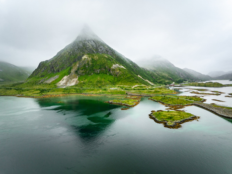 Aerial view of Volandstinden mountain with lush green slopes and misty peaks rising above the turquoise waters and rocky islands of the Lofoten archipelago in Lofoten Nordland Norway. Aerial view of Volandstinden mountain with lush green slopes and misty peaks rising above the turquoise waters and rocky islands of the Lofoten archipelago in Lofoten Nordland Norway.