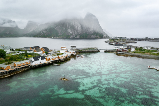 Aerial view of the fishing village of Reine with red and white wooden houses on stilts over turquoise fjord waters set against dramatic mist covered mountains in Reine Nordland Norway. Aerial view of the fishing village of Reine with red and white wooden houses on stilts over turquoise fjord waters set against dramatic mist covered mountains in Reine Nordland Norway.