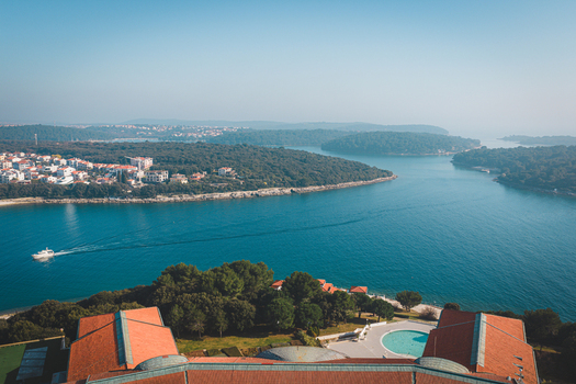 Aerial view of the rugged coastline where deep blue waters meet lush green islands and the ancient city a boat cutting through the shimmering surface Pula Istria County Croatia.