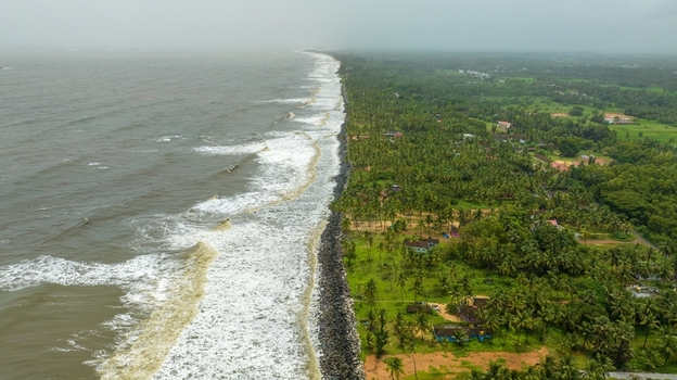 Aerial view of Kapu Light House standing along the lush green coastline with dense palm trees and crashing waves on the rocky shore in Udupi Karnataka India.
