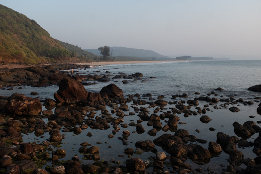 View of dark rugged rocks scattered along the shore meeting the gentle waves under a hazy sky with a backdrop of lush green hills Chiplun Maharashtra India. View of dark rugged rocks scattered along the shore meeting the gentle waves under a hazy sky with a backdrop of lush green hills Chiplun Maharashtra India.
