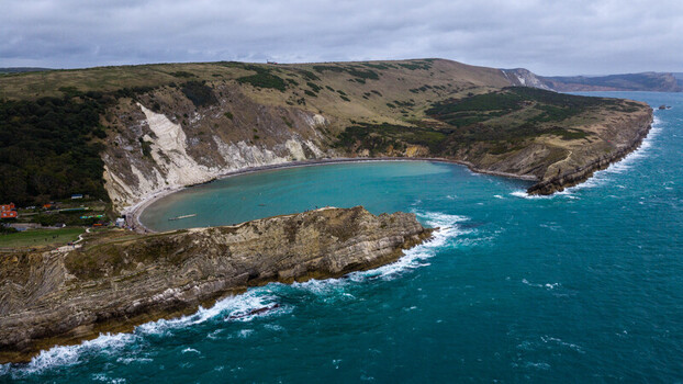 Aerial view of Lulworth Cove a horseshoe shaped bay with turquoise water dramatic limestone cliffs and grassy hills under a cloudy sky in Wareham England United Kingdom. Aerial view of Lulworth Cove a horseshoe shaped bay with turquoise water dramatic limestone cliffs and grassy hills under a cloudy sky in Wareham England United Kingdom.