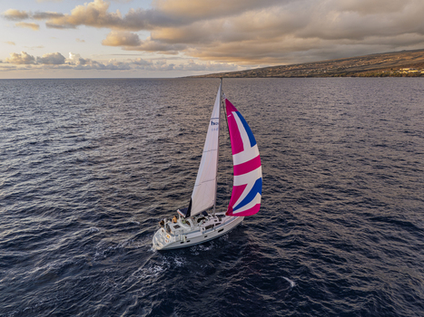 Aerial view of a sailboat with people on deck and a colorful spinnaker sailing on the deep blue ocean near the rugged coastline under a golden hour sky in Waimea Hawaii United States. Aerial view of a sailboat with people on deck and a colorful spinnaker sailing on the deep blue ocean near the rugged coastline under a golden hour sky in Waimea Hawaii United States.