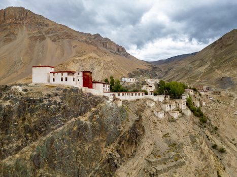 Aerial view of the Zanskar monastery perched on a rugged cliffside with white buildings and red accents surrounded by dramatic mountains under a cloudy sky in Zanskar Ladakh India.