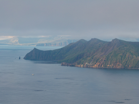 Aerial view of the rugged coastline of Unalaska with steep green mountains meeting the calm sea under a cloudy sky in Unalaska Alaska United States.