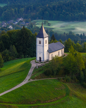 Aerial view of the Church of Saints Primus and Felician standing on a grassy hill surrounded by lush forests and rolling hills in the morning light in Jamnik Kranj Slovenia.