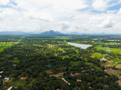 Aerial view of lush green forests scattered houses and a lake with mountains in the distance under a cloudy sky Sigiriya Central Province Sri Lanka. Aerial view of lush green forests scattered houses and a lake with mountains in the distance under a cloudy sky Sigiriya Central Province Sri Lanka.
