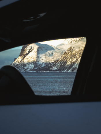 View of snow blanketed mountains meet the serene waters framed by the dark interior of a car creating a striking contrast of warmth and cold Indregård Troms Norway. View of snow blanketed mountains meet the serene waters framed by the dark interior of a car creating a striking contrast of warmth and cold Indregård Troms Norway.