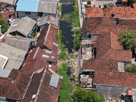 Aerial view of a dense residential neighborhood with terracotta tiled rooftops and a narrow canal flowing through the center in Kecamatan Denpasar Barat Bali Indonesia. Aerial view of a dense residential neighborhood with terracotta tiled rooftops and a narrow canal flowing through the center in Kecamatan Denpasar Barat Bali Indonesia.