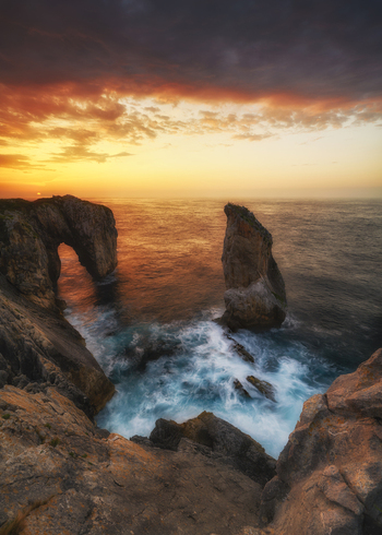 View of a natural rock arch and sea stack on a rugged coastline with crashing waves under a dramatic sunset sky Asturias Spain. View of a natural rock arch and sea stack on a rugged coastline with crashing waves under a dramatic sunset sky Asturias Spain.
