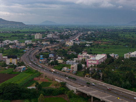 Aerial view of a highway winding through the town of Karad with surrounding green agricultural fields buildings and distant hills under a cloudy sky in Karad Maharashtra India.