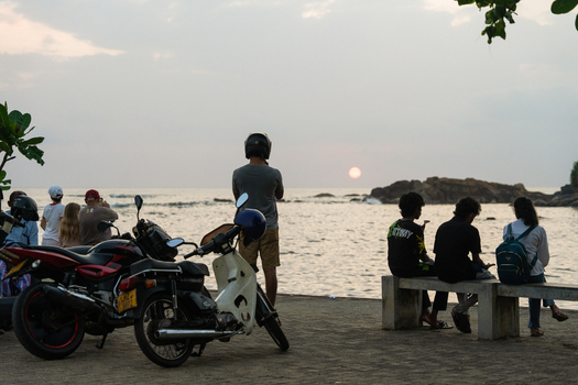 Sri Lanka 15 January 2026 View of people gathered on the coast to watch the sunset over the ocean with parked motorcycles and rocky formations.