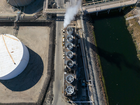Aerial view of an industrial cooling tower bank with steam rising adjacent to a large white storage tank pipelines and a waterway in Carson California United States.