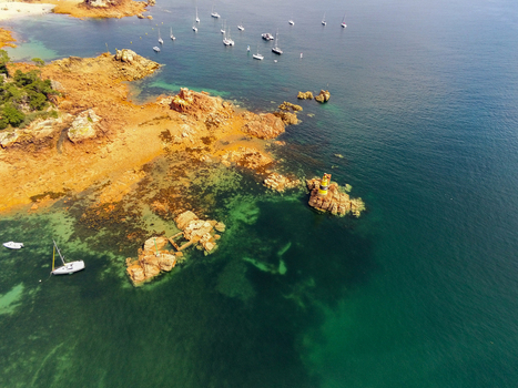Aerial view of the lighthouse at Brehat surrounded by rocky orange cliffs clear turquoise waters and several sailboats anchored in the bay in Ile de Brehat Brittany France.