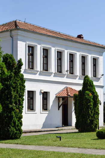 View of a historic white building with a red tiled roof framed by tall green cypress trees and a manicured lawn under a clear blue sky in Pristina Kosovo.