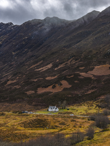 Aerial view of a small white cottage nestled at the foot of rugged misty mountains under a dramatic cloudy sky in Scotland United Kingdom. Aerial view of a small white cottage nestled at the foot of rugged misty mountains under a dramatic cloudy sky in Scotland United Kingdom.