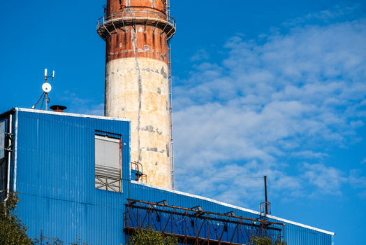 View of a tall weathered industrial chimney rising above a blue corrugated metal building under a bright blue sky in Poland.