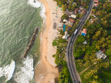 Aerial view of a sandy beach meeting turquoise waves and a coastal road lined with lush palm trees and small houses in Galle Southern Province Sri Lanka.