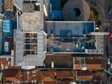 Aerial view of a modern building rooftop featuring industrial HVAC equipment and pipes situated next to a street with traffic and traditional tiled roof houses in Purwokerto Central Java Indonesia. Aerial view of a modern building rooftop featuring industrial HVAC equipment and pipes situated next to a street with traffic and traditional tiled roof houses in Purwokerto Central Java Indonesia.