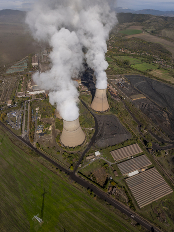 Aerial view of a coal fired power plant with large cooling towers emitting thick white steam situated in a rural landscape with industrial facilities and green fields in Bulgaria.