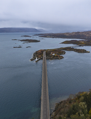 Aerial view of Skye Bridge spanning across the sea with a small lighthouse and rugged islands under a cloudy sky Isle of Skye Scotland United Kingdom.