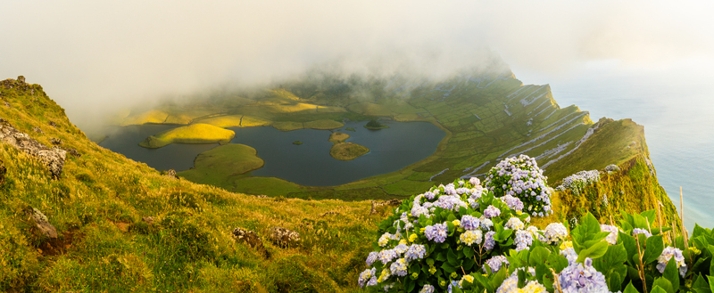 Aerial view of the Caldeirao volcanic crater lake on Corvo Island with lush green slopes blooming hydrangea flowers and mist rolling over the caldera in Vila do Corvo Azores Portugal.
