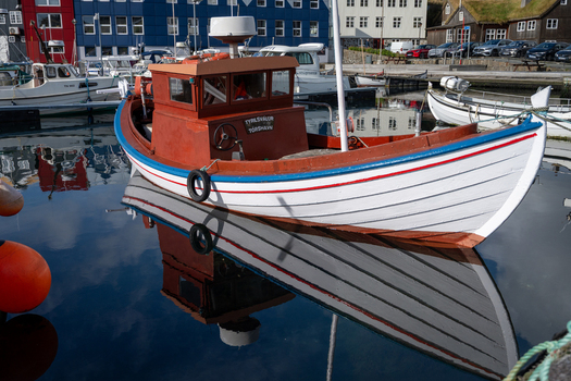 View of a traditional wooden fishing boat moored in the calm harbor waters with colorful buildings in the background in the Faroe Islands. View of a traditional wooden fishing boat moored in the calm harbor waters with colorful buildings in the background in the Faroe Islands.