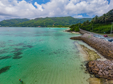 Aerial view of turquoise waters meeting the rocky shore cars parked along the coastal road against a backdrop of lush green hills Anse Royale Mahe Island Seychelles. Aerial view of turquoise waters meeting the rocky shore cars parked along the coastal road against a backdrop of lush green hills Anse Royale Mahe Island Seychelles.