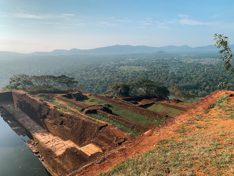 View of the ancient ruins and water tank atop the Sigiriya rock fortress overlooking the vast lush jungle and distant mountains in Sri Lanka.