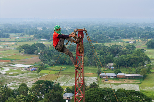 Trishal Upazila Bangladesh 13 May 2026 Aerial view of a worker climbing a tall red metal tower overlooking lush green agricultural fields and rural landscapes. Trishal Upazila Bangladesh 13 May 2026 Aerial view of a worker climbing a tall red metal tower overlooking lush green agricultural fields and rural landscapes.