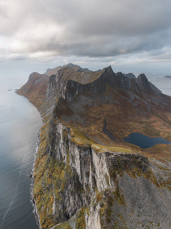 Aerial view of the dramatic Segla mountain ridge on Senja island featuring steep rocky cliffs autumn colored vegetation and a small alpine lake in Senja Troms Norway.