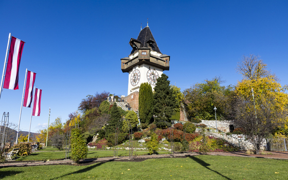 View of the iconic Uhrturm clock tower perched atop a hill surrounded by vibrant autumn foliage and flags waving gently in the breeze Graz Styria Austria. View of the iconic Uhrturm clock tower perched atop a hill surrounded by vibrant autumn foliage and flags waving gently in the breeze Graz Styria Austria.