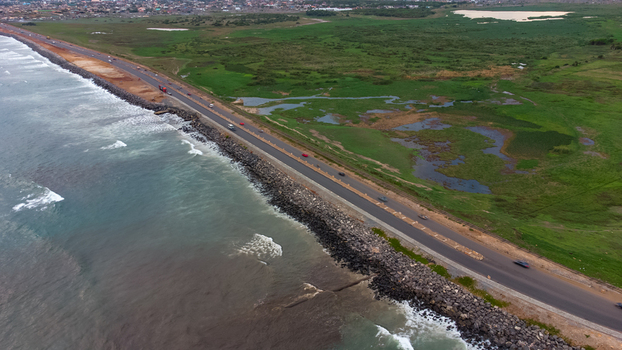 Aerial view of the Accra Tema Beach Road stretching along the coastline with crashing waves rocky embankments and adjacent green wetlands Accra Greater Accra Region Ghana.