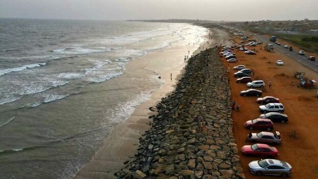 Aerial view of a coastline with a stone seawall separating the sandy beach from a parking area filled with cars along the road in Ghana. Aerial view of a coastline with a stone seawall separating the sandy beach from a parking area filled with cars along the road in Ghana.