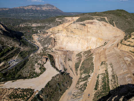 Aerial view of a large open pit quarry with terraced rock faces dusty roads and heavy machinery in a mountainous landscape near Teulada Valencian Community Spain.