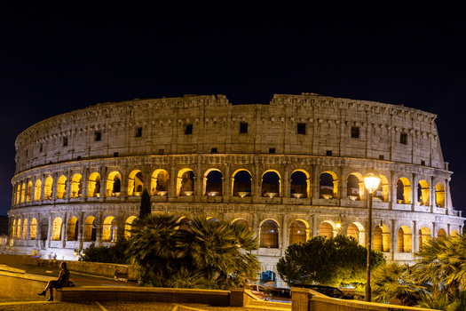 View of the illuminated Colosseum at night with palm trees and a person sitting on a wall in the foreground Roma Lazio Italy.